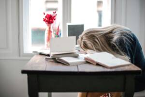 a girl with her head on a brown table which also has a laptop, vase and books.
